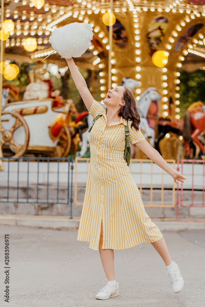 Obraz premium Young woman with cotton candy in front of the carrousel with night illumination at the amusement park.