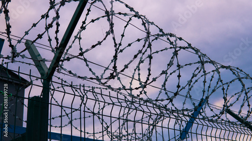 The fence of the correctional facility with barbed wire on the background of a gloomy cloudy dark blue sky.