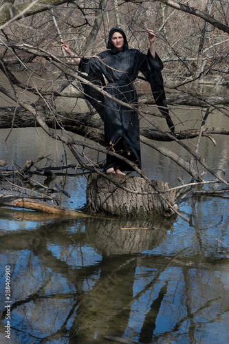 Photos Hooded woman in black robes standing on a tree stump in the middle of a swamp