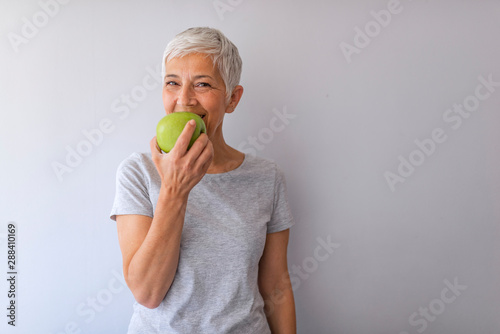 Portrait of happy mature woman holding granny smith apple at home. Beautiful senior woman over grunge grey wall eating green apple with happy face smiling.