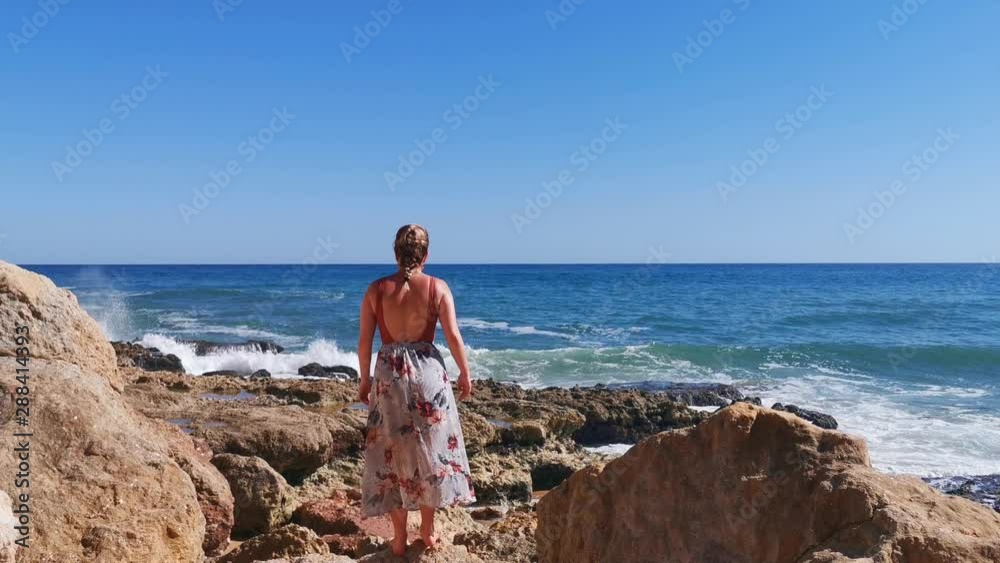Static shot, of a woman walking on rocks, in a dress, looking at waves hitting the coast of praia dos Caneiros, at the Atlantic sea, on a sunny summer day, in Lagos, Algarve, Portugal Static shot, of a woman walking on rocks, in a dress, looking at waves hitting the coast of praia dos Caneiros, at the Atlantic sea, on a sunny summer day, in Lagos, Algarve, Portugal