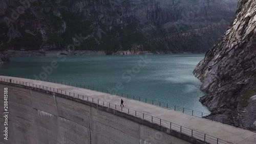 Aerial view of the Limmernsee reservoir and dam near Linthal (Switzerland) while someone is walking on the dam. The drone is flying backward from the dam. The mountains can be seen in the background. photo