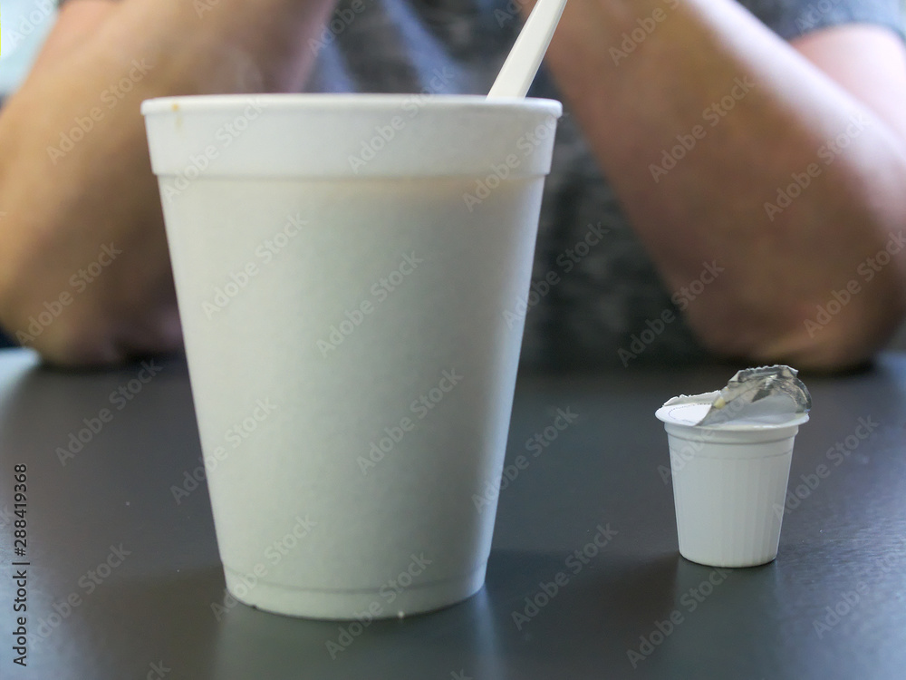 Styrofoam coffee cup with plastic spoon and used creamer container in  foreground and human elbows on table in background. Stock Photo