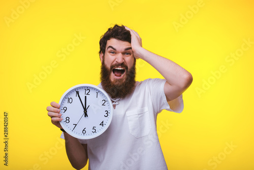 A handsome upset guy, holding a clock over yellow background