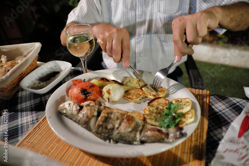 Dinner at the restaurant. Mediterranean food. Grilled fish and vegetables on a plate. Hands of a man cutting fish in a plate.