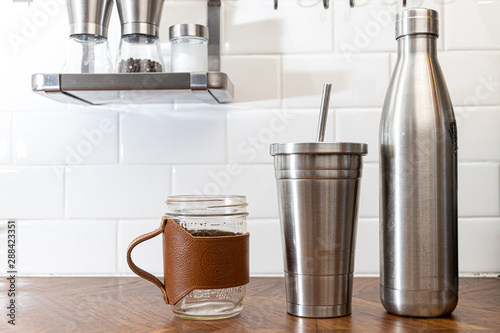 Reusable eco friendly stainless steel drinking and glass mugs and bottles for a range of beverages in a modern kitchen with wood counters and white subway tile wall
