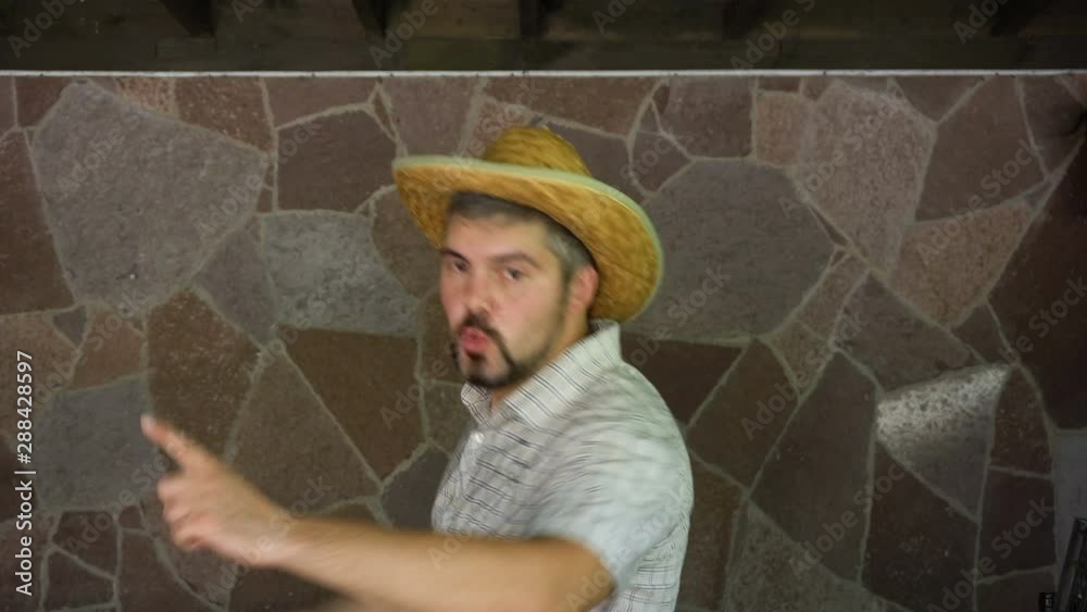 A man in a shirt and a straw hat is dancing against a stone wall. On