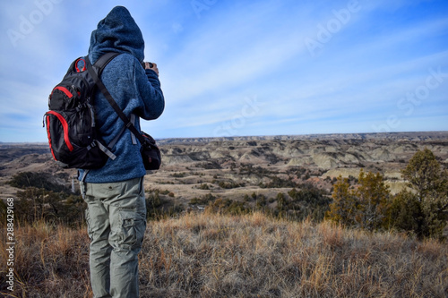 The back side of a hiker wearing a blue hoodie, khaki cargo pants, red and black backpack with a satchel taking a photo of the beautiful scenic landscape ahead with rolling hills and canyons.