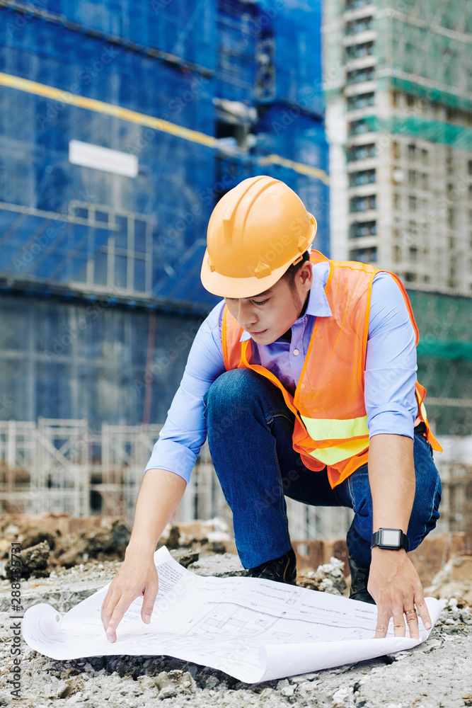 Professional Asian civil engineer spreading out building blueprint on the ground and examining floor plan Professional Asian civil engineer spreading out building blueprint on the ground and examining floor plan
