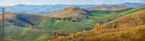 Autumn panoramic view. Hilly terrain. Yellow slopes and trees contrast with green fields.