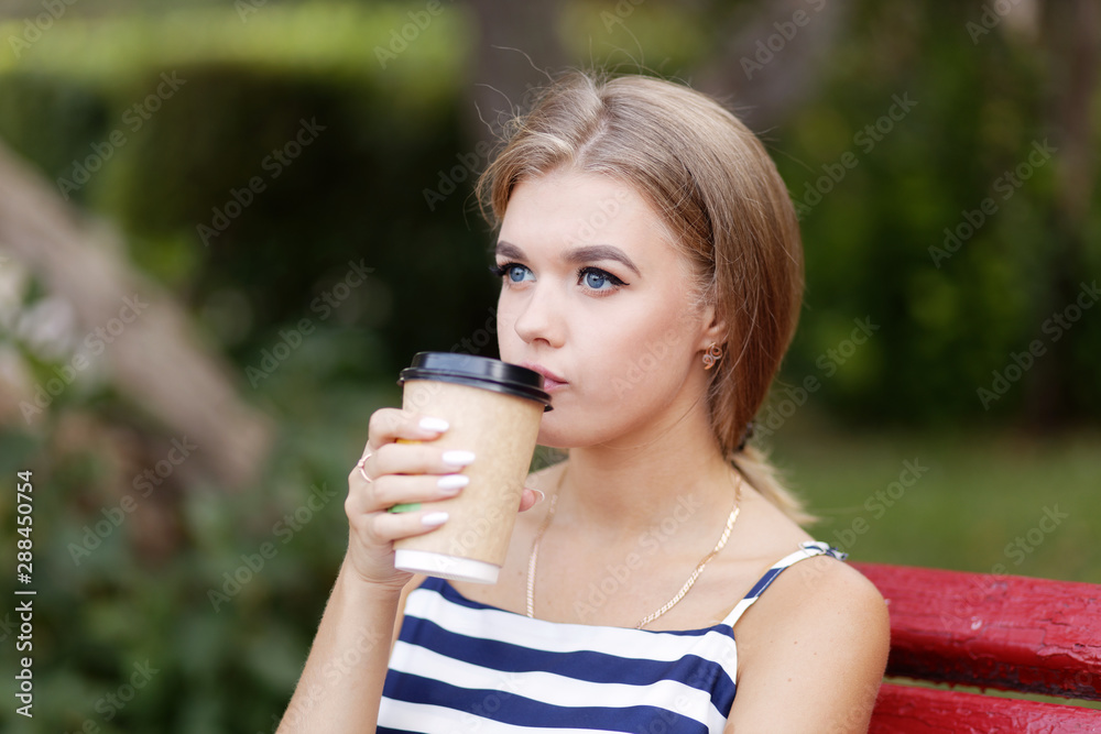 Young girl sitting alone on a park bench and drinking coffee from a cardboard cup. Shallow focus.