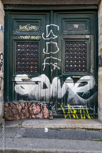 Old wood door painted in dark green color and damaged by colorful paints. Vintage door with ornate grids. Doorway in dangerous district 11 of Paris. Cultural contrast. Paris, France - June 26, 2019 photo