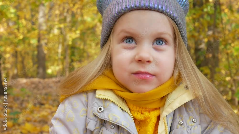 Little girl in neon hat and orange scarf eats something and smiles in forest or park on background of yellow foliage in fall Little girl in neon hat and orange scarf eats something and smiles in forest or park on background of yellow foliage in fall