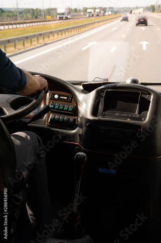 A Caucasian male driving an old bus along highway, asphalt road surface illuminated by bright daylight, cars seen in the horizon.