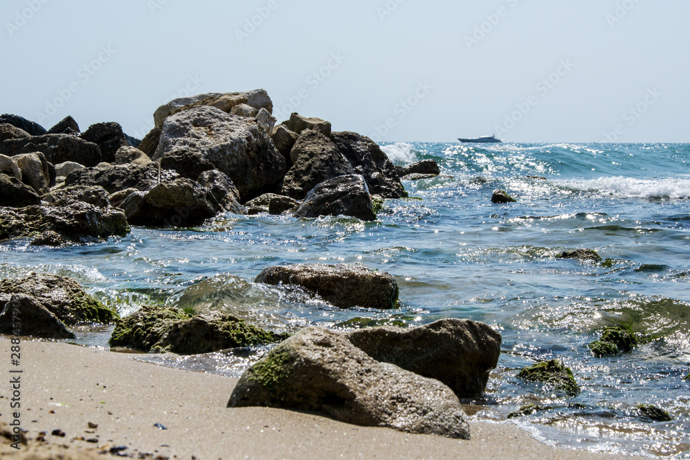 Beautiful wild beach landscape, sunny day, water waves hitting the cliffs, nature summertime scene, blurred yacht in the background