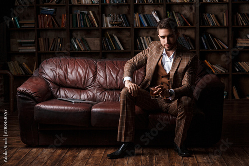 young successful man in a brown kosmtyum drinks whiskey. bearded businessman sitting in a library on a luxurious leather sofa and drinking cognac