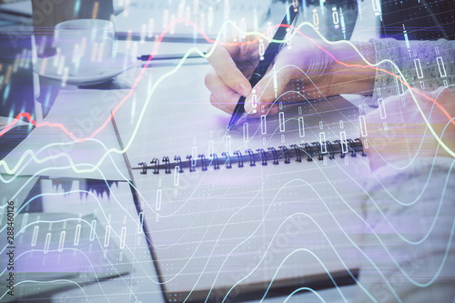 Hand taking notes in notepad. Forex chart holograms in front. Concept of research. Double exposure