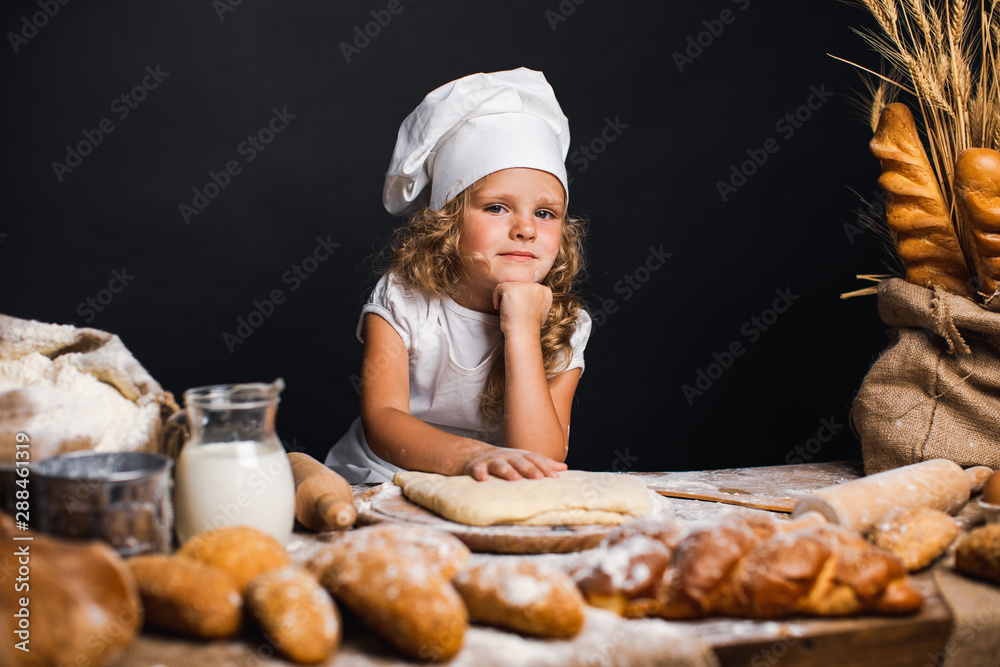 Charming little girl with curly hair in white apron and hat standing at table kneading bread dough and looking at camera. KId in good mood, having fun. Charming little girl with curly hair in white apron and hat standing at table kneading bread dough and looking at camera. KId in good mood, having fun.