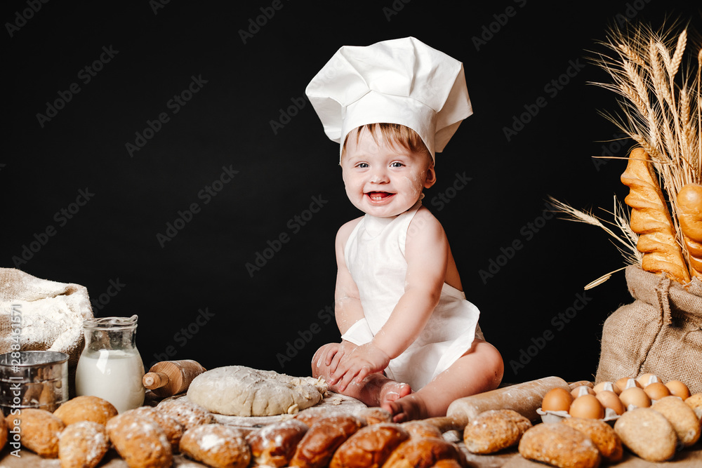 Charming toddler baby in hat of cook and apron sitting on table with bread loaves and cooking ingredients laughing happily Charming toddler baby in hat of cook and apron sitting on table with bread loaves and cooking ingredients laughing happily