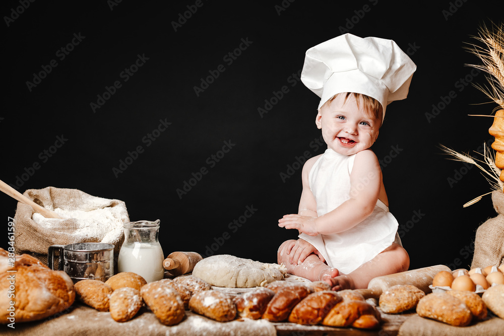 Charming toddler baby in hat of cook and apron sitting on table with bread loaves and cooking ingredients laughing happily Charming toddler baby in hat of cook and apron sitting on table with bread loaves and cooking ingredients laughing happily