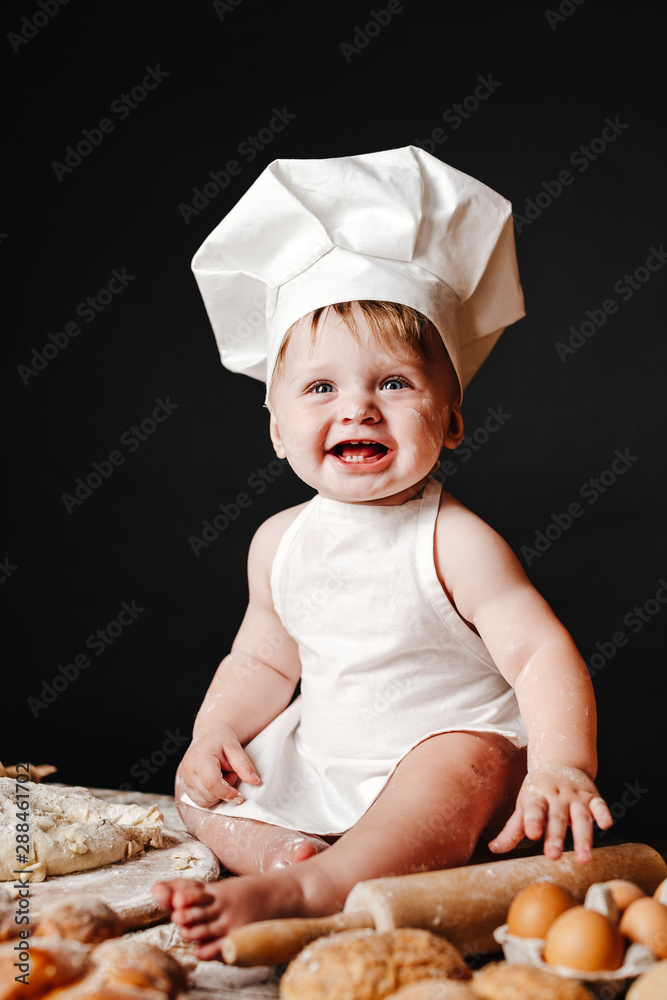 Charming toddler baby in hat of cook and apron sitting on table with bread loaves and cooking ingredients laughing happily Charming toddler baby in hat of cook and apron sitting on table with bread loaves and cooking ingredients laughing happily