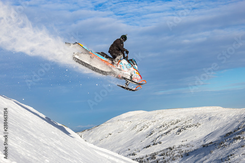 a snowmobile flies in the mountains with a large snow trail against a bright blue sky with clouds. Snowmobile jump at high altitude. bright snowmobile and looking at the camera. super quality photo