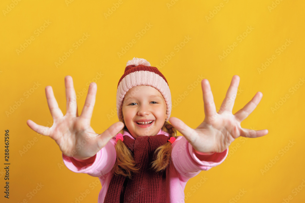 Cheerful little girl in a scarf and hat on a yellow background. The child shows two palms, give five Cheerful little girl in a scarf and hat on a yellow background. The child shows two palms, give five