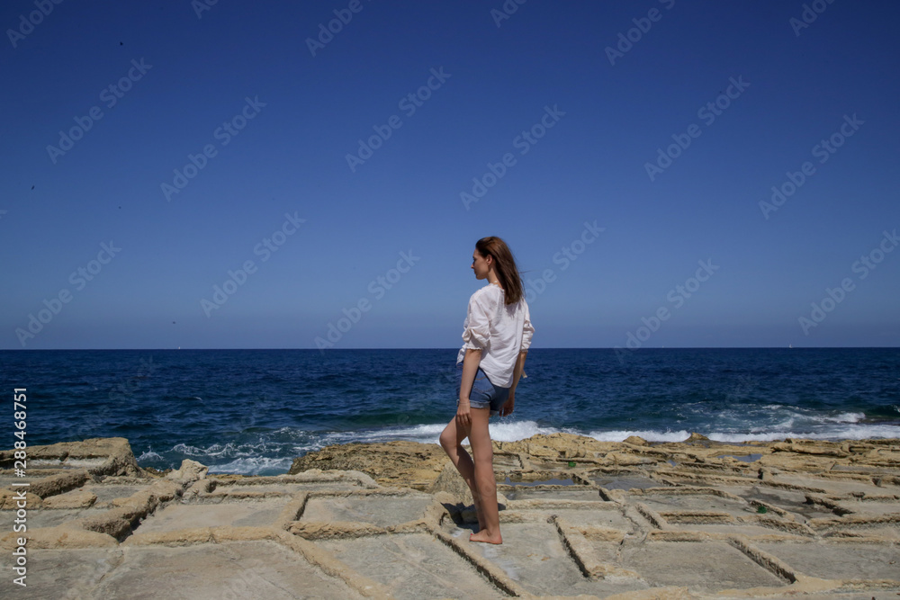 Young pretty woman in a white linen shirt and denim shorts on the windy rocky coast, enjoying summer vacation on the remote location far away from the tourist attractions