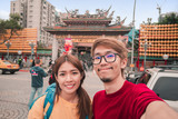 Asian man and woman traveler with backpack standing take photo selfie at Longshan Buddhist temple, landmark temple in taiwan.