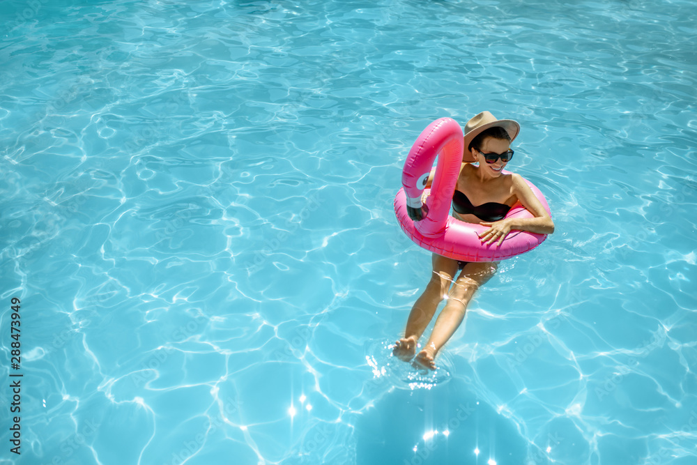 Woman in swimsuit and hat swimming with inflatable flamingo ring in the water pool, enjoying vacations during the summer time