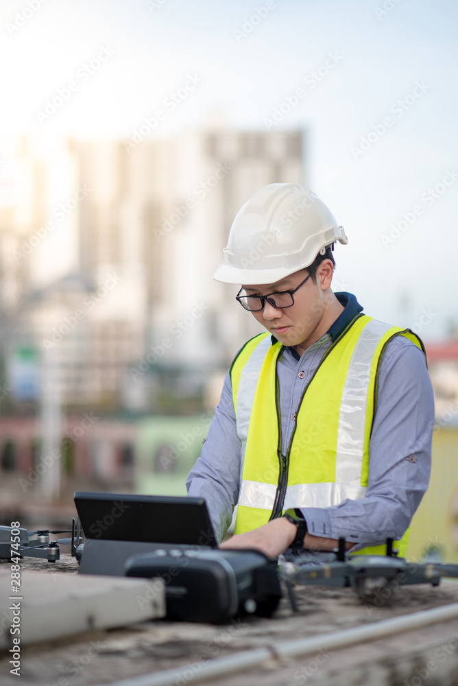 Asian engineer man working with drone, laptop and working tools at construction site. Male worker using unmanned aerial vehicle (UAV) for land and building site survey in civil engineering project.