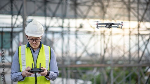 Asian engineer man flying drone over construction site. Male worker using unmanned aerial vehicle (UAV) for land and building site survey in civil engineering project.