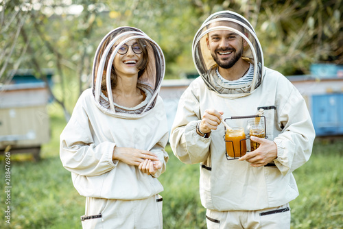 Portrait of a man and woman beekepers in protective uniform standing together with honey in the jar, tasting fresh product on the apiary outdoors photo