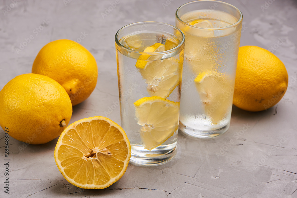 Glasses filled with pure water with lemon slices surrounded by a few whole lemon on the gray concrete background. Close up Glasses filled with pure water with lemon slices surrounded by a few whole lemon on the gray concrete background. Close up