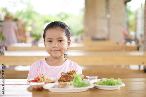 Asian child cute with dimple or kid girl enjoy eating grilled chicken and papaya salad for Thai food and appetizing on table with smiling and happy for breakfast or lunch in restaurant or food court