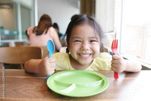 Asian child cute hungry or kid girl holding colorful spoon and fork with dish for wait eat delicious food with smiling and happy for enjoy lunch or breakfast in morning at restaurant or food court photo