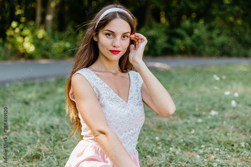 Exquisite, charming, sexy, beautiful brunette girl in blouse and headband poses sitting on green lawn in summer park Exquisite, charming, sexy, beautiful brunette girl in blouse and headband poses sitting on green lawn in summer park
