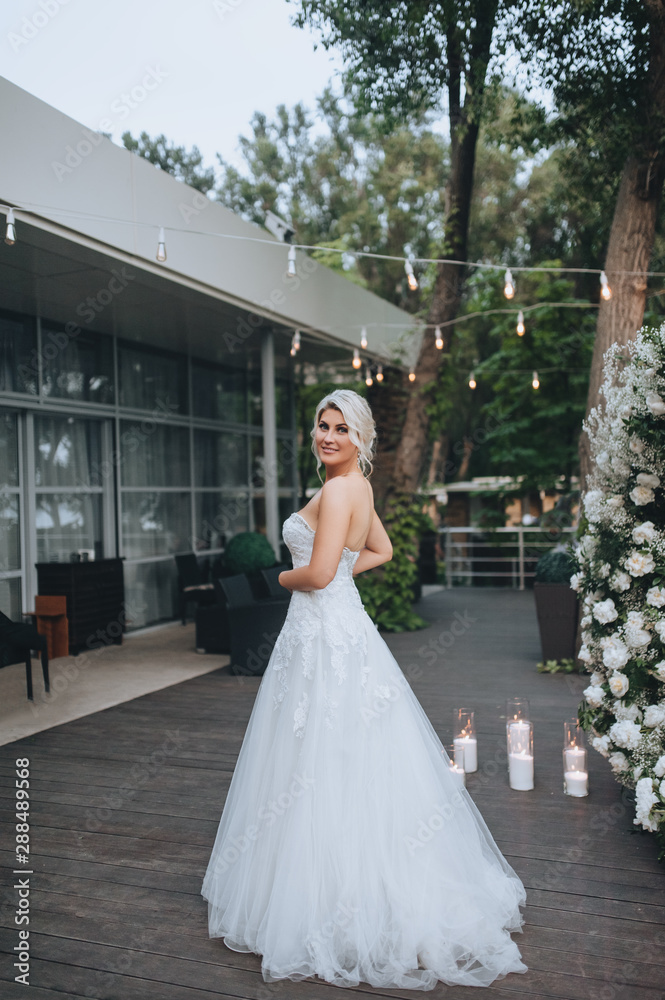 Beautiful blonde bride dancing and smiling in a restaurant against the background of garlands and light bulbs. Wedding portrait of a cute girl in a white dress. Concept and photography.