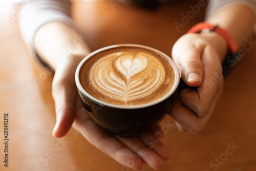 Close up Cup of coffee latte in coffee shop.Female hands holding a cup of coffee cup with heart shaped latte art foam on black wood table.