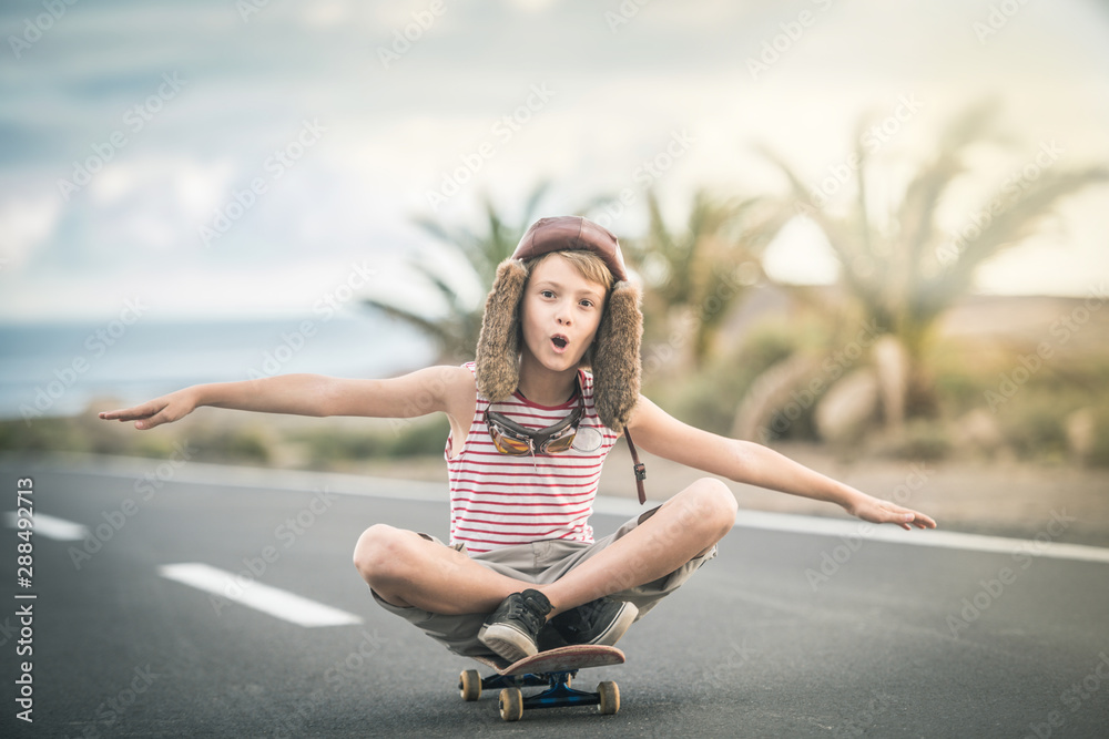 Young blond boy plays aviator sitting cross-legged on skateboard with arms outstretched to fly smiling child imitates plane flying on airport runway. Concept image of a plane taking off for a vacation