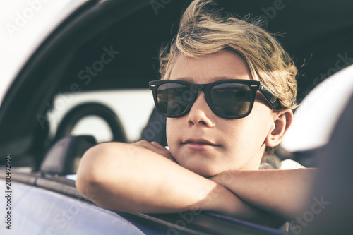 Portrait of beautiful boy looking out of the car window. Close up of a caucasian teen with sunglasses and arms crossed. Cool teenager wind in the hair looking away. Youth, positive, freedom concept.