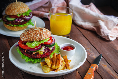 healthy vegetarian burger with beet patty coleslaw salad fresh vegetables fries potato and bowl with tomato sauce glass of orange  juice knife on wooden table