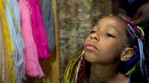 African hairdresser plaits braids with multicolored artificial strands for mixed Jamaica ethnicity little girl. Portrait of a girl with curly long hair