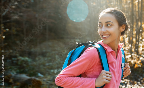 portrait of woman hiker with small backpack, wearing in pink fleece jacket, standing on pine woods background in the Carpathians woody mountains, Western Ukraine. happy smiling woman looking back at photo