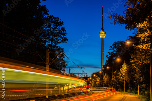 Germany, Fast motion of driving train alongside traffic road with illuminated tv tower building behind in magical twilight mood after sunset in summer photo