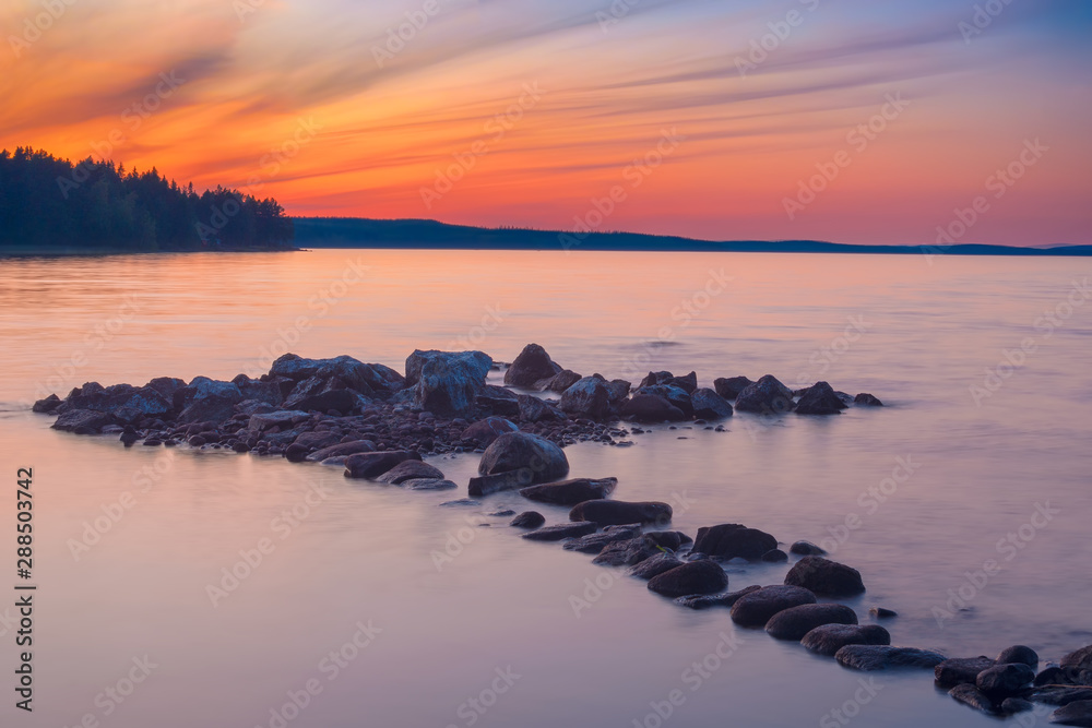 Long Exposure of Sunset at the Paijanne lake. Beautiful scape with stone beach, pine forest and water. Lake Paijanne, Finland. Long Exposure of Sunset at the Paijanne lake. Beautiful scape with stone beach, pine forest and water. Lake Paijanne, Finland.