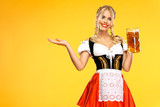 Young sexy Oktoberfest girl waitress, wearing a traditional Bavarian or german dirndl, holding big beer mug with drink isolated on yellow background. Woman pointing to looking left.