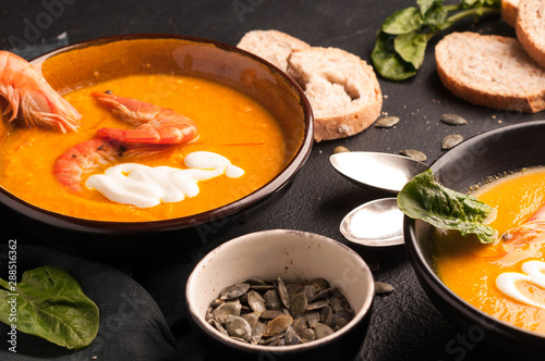 side view of pumpkin soup with prawns and sour in bowls near homemade bread, seeds, herbs on black background