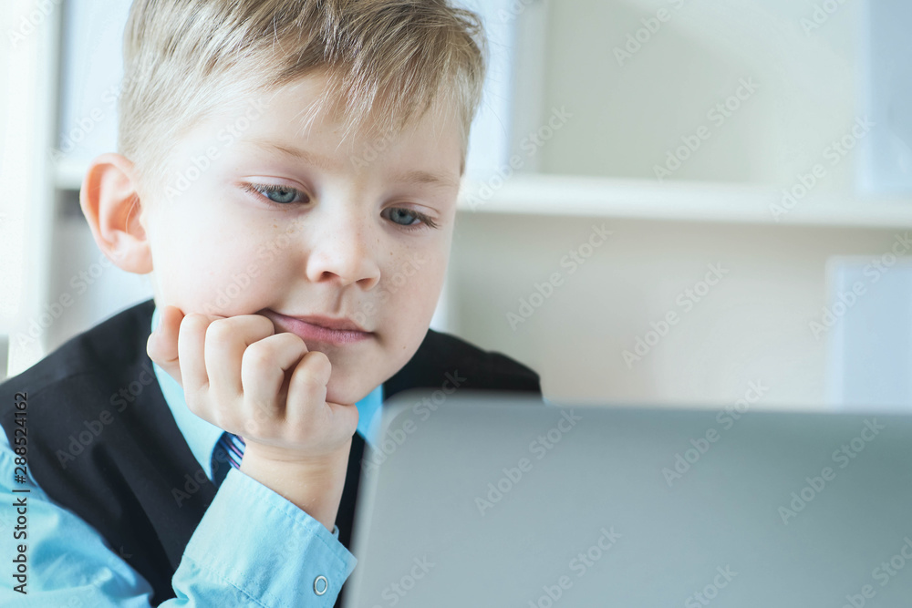 Small cute boy in business suit works at computer at office and smiling. Small business man, little boss. Small cute boy in business suit works at computer at office and smiling. Small business man, little boss.
