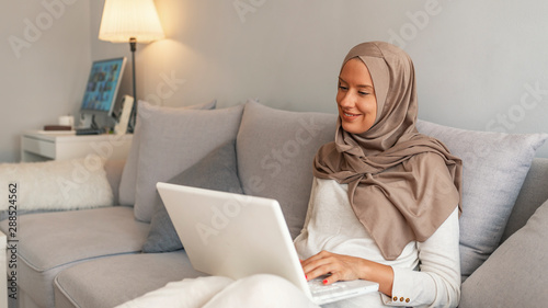 Muslim woman Using her Laptop, Surfing Social Networks at Home, Free Space. Surfing the net at home. Side view of attractive young Muslim woman working on laptop and smiling